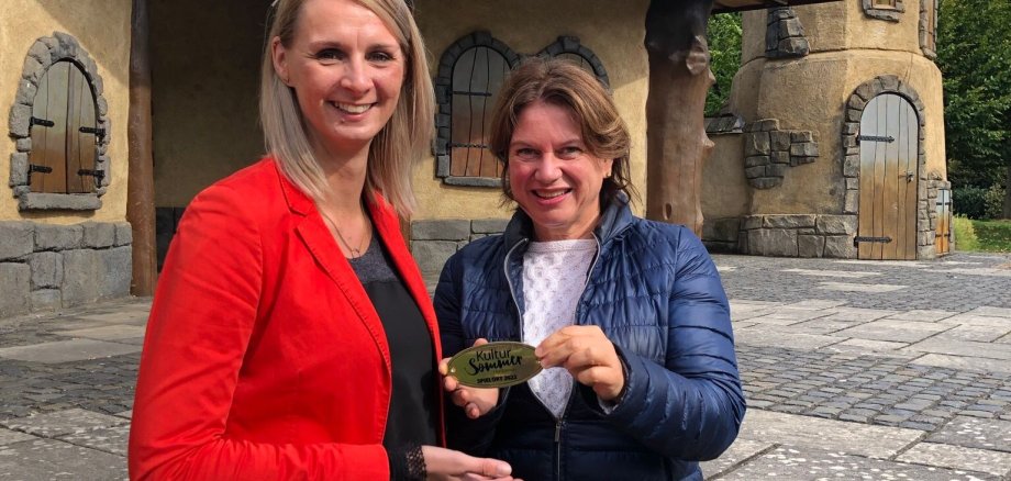 Bürgermeisterin Sina Best (links) bekommt von Frau Matthes (Geschäftsführerin vom Kultursommer Nordhessen) die goldene Plakette „Spielort 2022“ für die Gudensberger Märchenbühne überreicht.