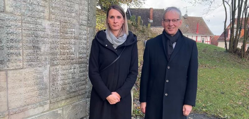 Mann und Frau vor Wand mit Namen von Gefallenen mit Kranz