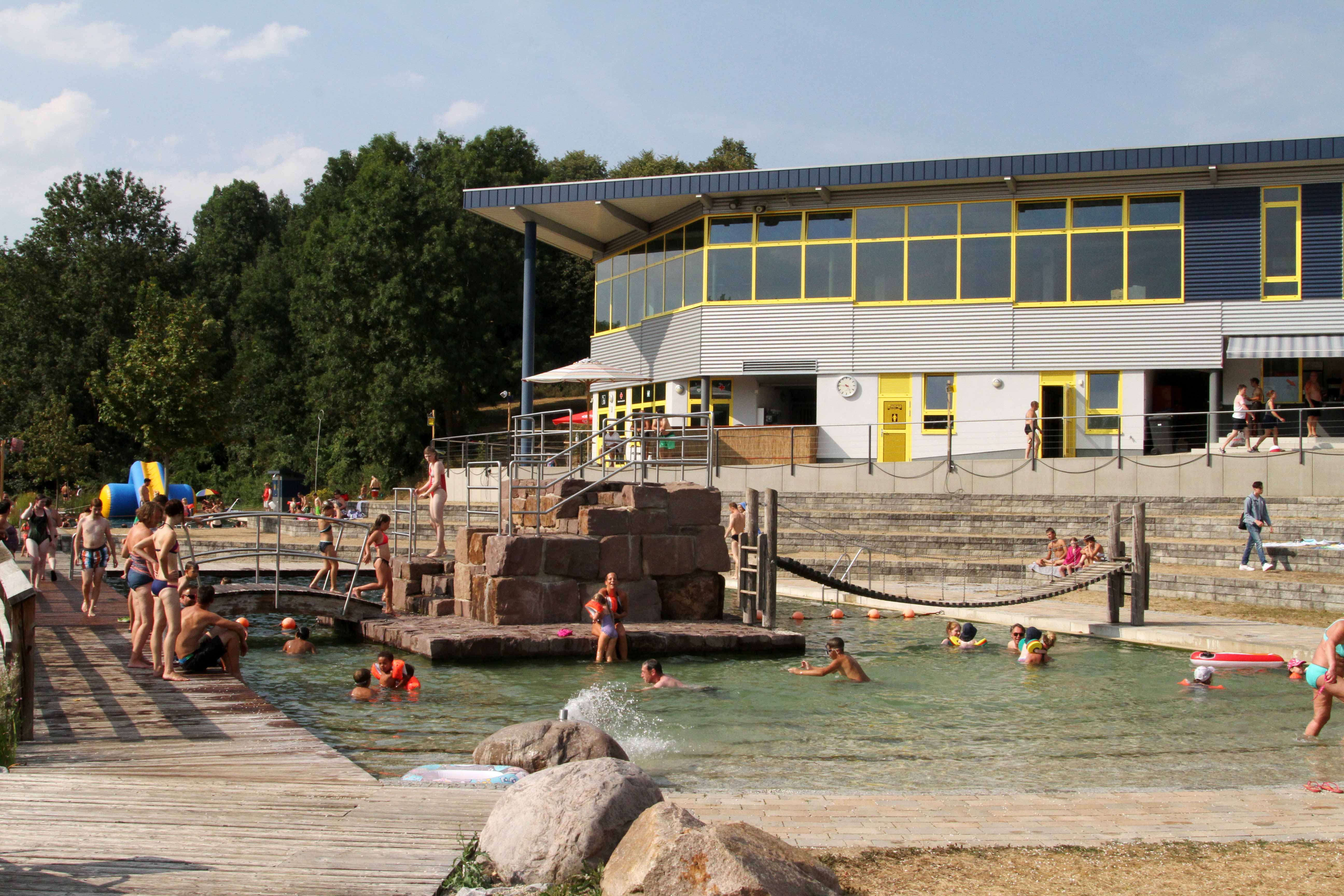 Schwimmbecken im Naturbad mit schwimmenden und danebensitzenden Menschen. 