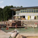 Schwimmbecken im Naturbad mit schwimmenden und danebensitzenden Menschen. 