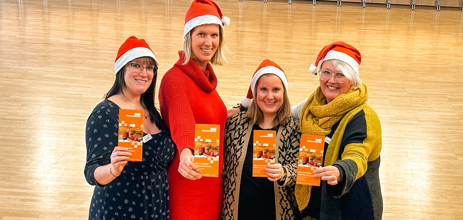 Vier Frauen mit Weihnachtsmannmützen halten Flyer vom Weihnachtsmarkt