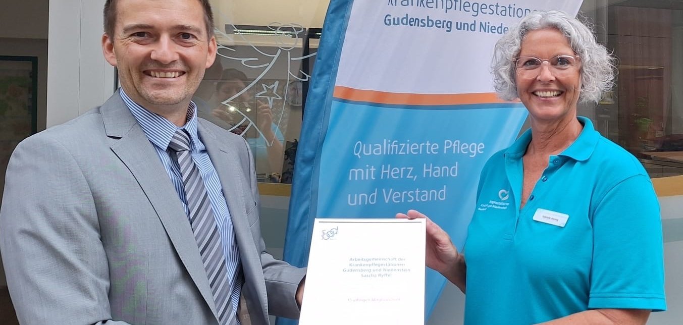 Ein Herr und die Leiterin der Krankenpflege vor einem Banner mit dem Zertifikat in der Hand