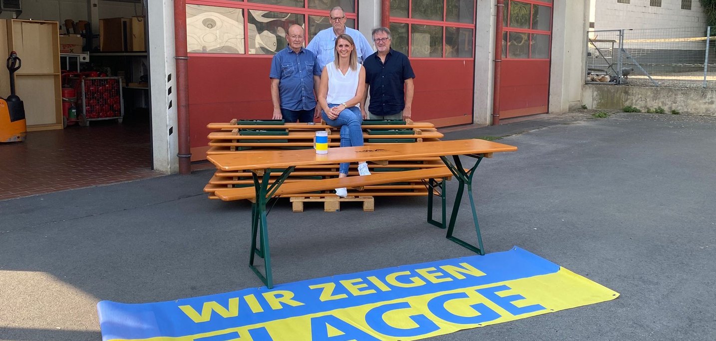Drei Männer und eine Frau auf Bierzeltgarnituren vor einer Feuerwehr. Davor eine Flagge in blau/gelb mit der Aufschrift: Wir zeigen Flagge