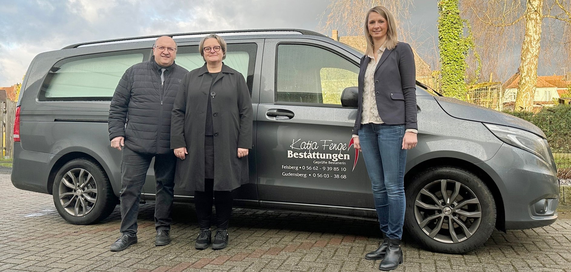 zwei Frauen und ein Mann vor einem Leichenwagen mit Aufschrift Bestattungsinstitut Hess