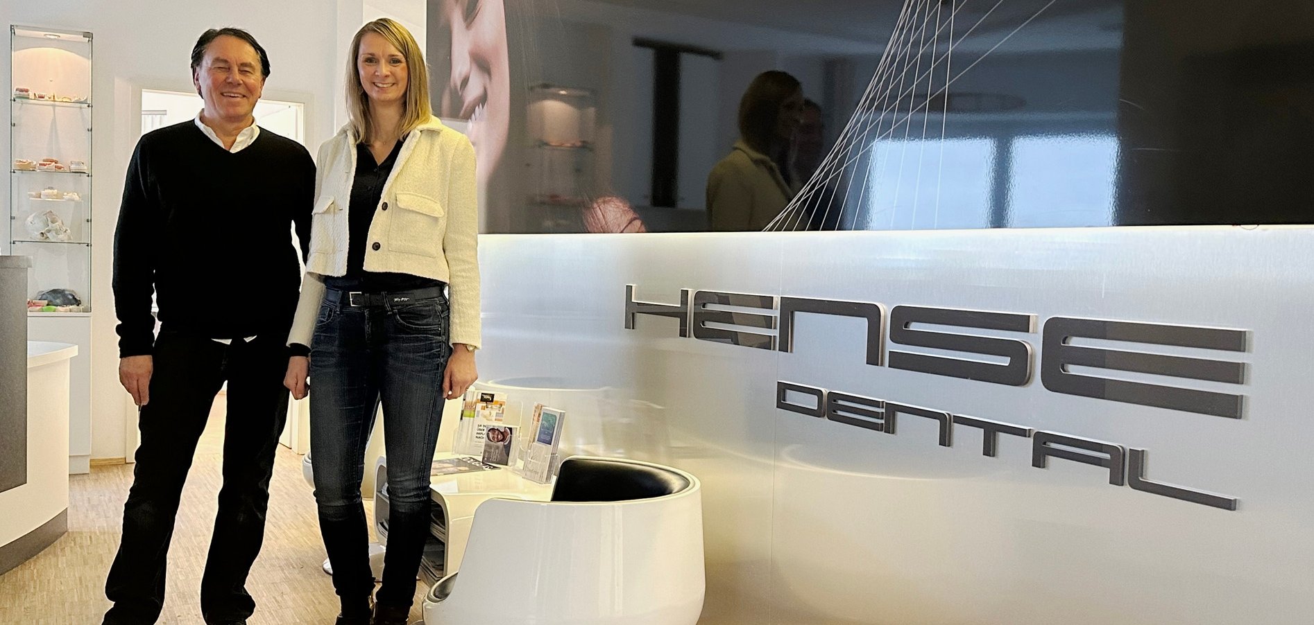 Mann und Frau im Flur mit Stühlen. An der Wand die Schrift: Hense Dental